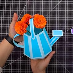 a person holding a blue watering can with orange flowers in it and an origami flower on top