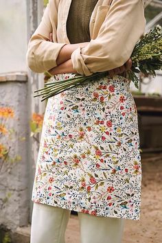 a woman standing with her arms crossed wearing a floral apron and holding flowers in her hands