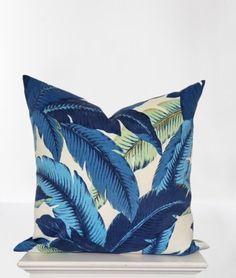 a blue and white pillow with green palm leaves on the front, sitting on a white pedestal