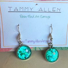 a pair of earrings with green and blue glitter on them sitting next to a card