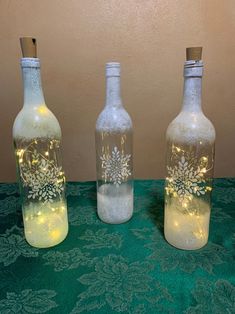 three bottles with lights in them sitting on a green tableclothed surface, one is empty and the other has snowflakes