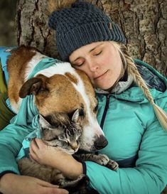 a woman holding a cat and dog in her arms while sitting next to a tree