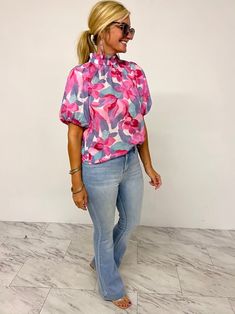 a woman standing in front of a white wall wearing jeans and a pink floral shirt