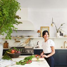 a woman standing in a kitchen preparing food on top of a wooden cutting board next to a potted plant