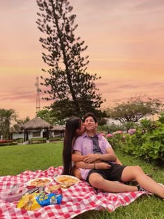 a man and woman sitting on a blanket in the grass near a picnic table with food