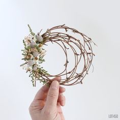 a hand holding a wreath with flowers and twigs on it's side, in front of a white background