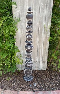 a tall metal sculpture sitting in front of a wooden fence next to a green bush