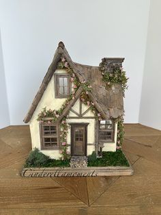 a small white house with flowers on the roof and windows, sitting on top of a wooden table