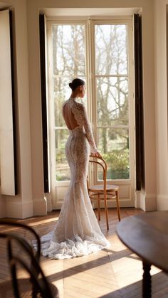 a woman standing in front of a window wearing a long sleeved wedding dress with an open back