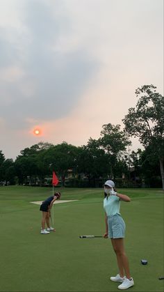 two women playing golf on a green field with trees in the background and sun setting behind them