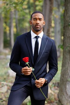 a man in a suit and tie holding a red rose while standing next to a tree