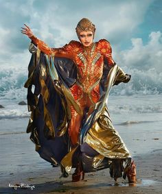 a woman in an orange and gold costume standing on the beach with her arms outstretched