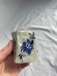 a hand holding a blue and white flowered square vase on a white sheet background