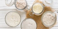 four glass jars filled with different types of flour on top of a wooden table next to a burlap bag