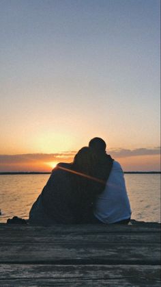 two people are sitting on a dock and hugging each other as the sun sets in the background