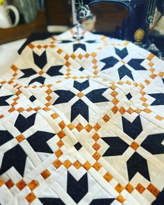 a quilted table topper with an orange and black star design on it, next to a sewing machine