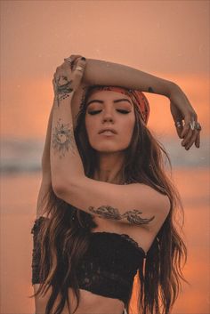 a woman with long hair and tattoos on her arm standing in front of the ocean