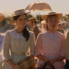 three women in dresses and hats are standing on the beach with umbrellas behind them