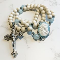 "This handcrafted, beautiful, and unique Catholic rosary is made with 8mm bleached white wood beads and 8mm aqua quartz beads. The beads are strung and knotted on micro cord (the slimmest type of paracord!) for durability. The cameo rosary center depicts the Madonna of the Streets. The crucifix is 2 inches in length and was made in Italy.  This rosary is about 14 inches in length and is lightweight and easy to carry with you so you can \"pray without ceasing.\" Each rosary from Refuge Rosaries is carefully packed in a cotton muslin drawstring bag that can be used as a rosary case. Follow us on Instagram and Facebook @refugerosaries to stay in touch and see our latest designs!" Rosary Case, Upcycled Vintage Jewelry, Rosary Beads Catholic, Pray Without Ceasing, Catholic Rosary, Rosary Catholic, Rosary Beads, Catholic Gifts, Quartz Beads