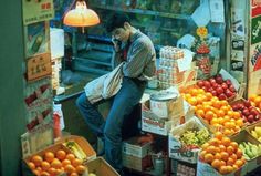 a man standing in front of a fruit stand talking on a cell phone while surrounded by boxes of oranges and other fruits