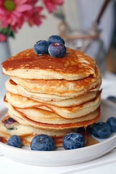 a stack of pancakes with blueberries and syrup on a white plate next to pink flowers