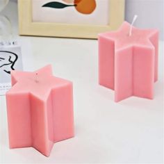 two pink candles sitting next to each other on a white table with a framed picture in the background