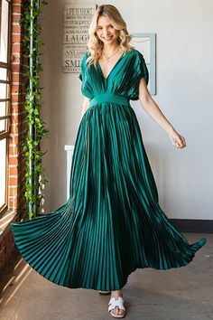 a woman wearing a green pleated dress standing in front of a brick wall and smiling at the camera