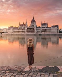 a woman standing on the edge of a lake looking at a large building in the background