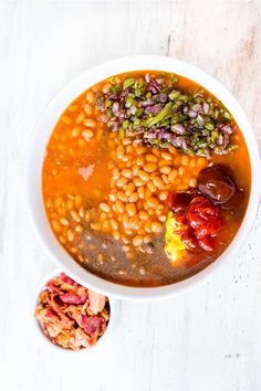 a white bowl filled with beans and other food on top of a wooden table next to a spoon