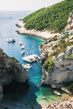 several boats are docked in the blue water near some cliffs and people on the beach