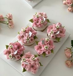 cupcakes decorated with pink flowers and green leaves on a white plate next to roses