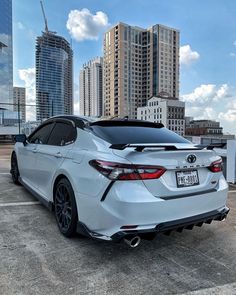 the rear end of a white car parked in a parking lot with tall buildings behind it