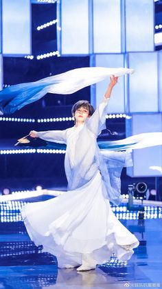 a woman in a white dress is dancing with her arms spread out and two blue umbrellas are flying above her head