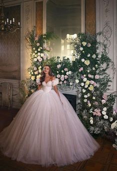 a woman in a ball gown standing next to a fireplace with flowers on the mantle