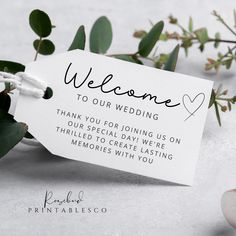 a wedding welcome card with flowers and greenery in the background, on top of a table