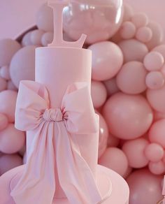 there is a pink cake with a bow on the top and balloons in the background