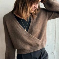 a woman with long hair wearing a brown sweater and black pants, leaning against a wall