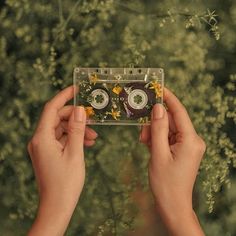 two hands holding up a cassette with eyes and flowers on it in front of trees