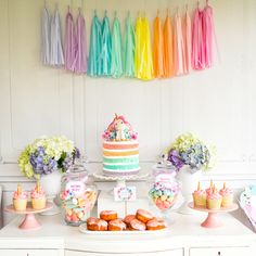 a table topped with lots of cakes and desserts next to rainbow streamers hanging from the ceiling