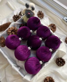 purple christmas ornaments are arranged on a white tablecloth with pine cones and small teddy bear