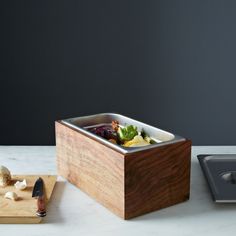 a wooden cutting board topped with food next to a container filled with fruit and vegetables