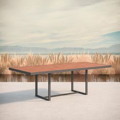 a wooden table with metal legs in front of a lake and reeds on the shore