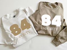 A sporty crew neck sweatshirt with the number 84 could be a nod to various things depending on the context. For example, it could represent a team number or a year that's significant to you. If it's for a sports team, it might be the jersey number of a famous player or just a number that's meaningful to the team. If it's more personal, maybe it's a favorite number or a reference to a special event or year. What's the story behind the number 84 for you? HOW TO ORDER 1) Select your choice of SIZE Collegiate White Top With Number Print, Collegiate White Tops With Number Print, White Sports T-shirt With Ribbed Cuffs, Sporty White Sweatshirt With Text Print, White Sporty Sweatshirt With Text Print, Sporty Logo Print Sweatshirt For Sports Events, White Number Print Top For Sports Season, Sporty White Tops With Number Print, Sporty White Top With Number Print