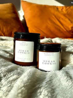 two jars of sugar cookie sitting on top of a white blanket next to an orange pillow