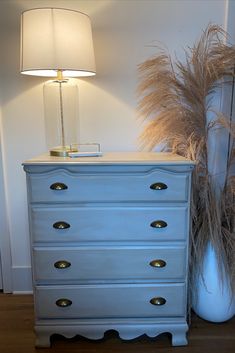 a blue chest of drawers next to a white vase with dried grass in it and a lamp on top