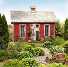 a small red house surrounded by greenery and flowers in the front yard, with a brick pathway leading to it