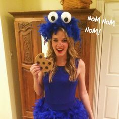 a girl in a blue dress is holding a cookie and wearing a cookie monster hat