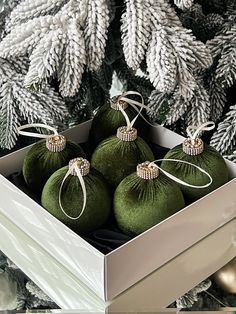 four green ornaments in a white box on a table next to christmas tree with pine cones