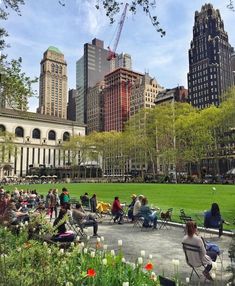 many people are sitting on benches in the middle of a park with tall buildings behind them