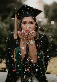 a woman wearing a graduation cap and gown blowing confetti in front of her face